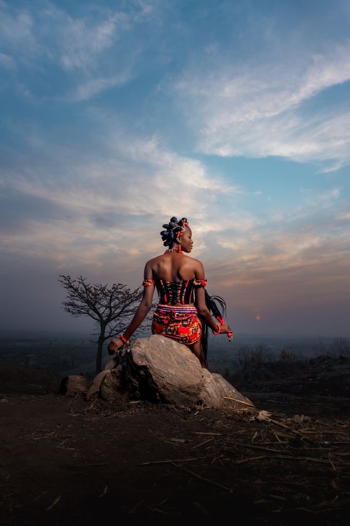 Back view of an African woman in traditional Nigerian attire facing a sunset landscape.