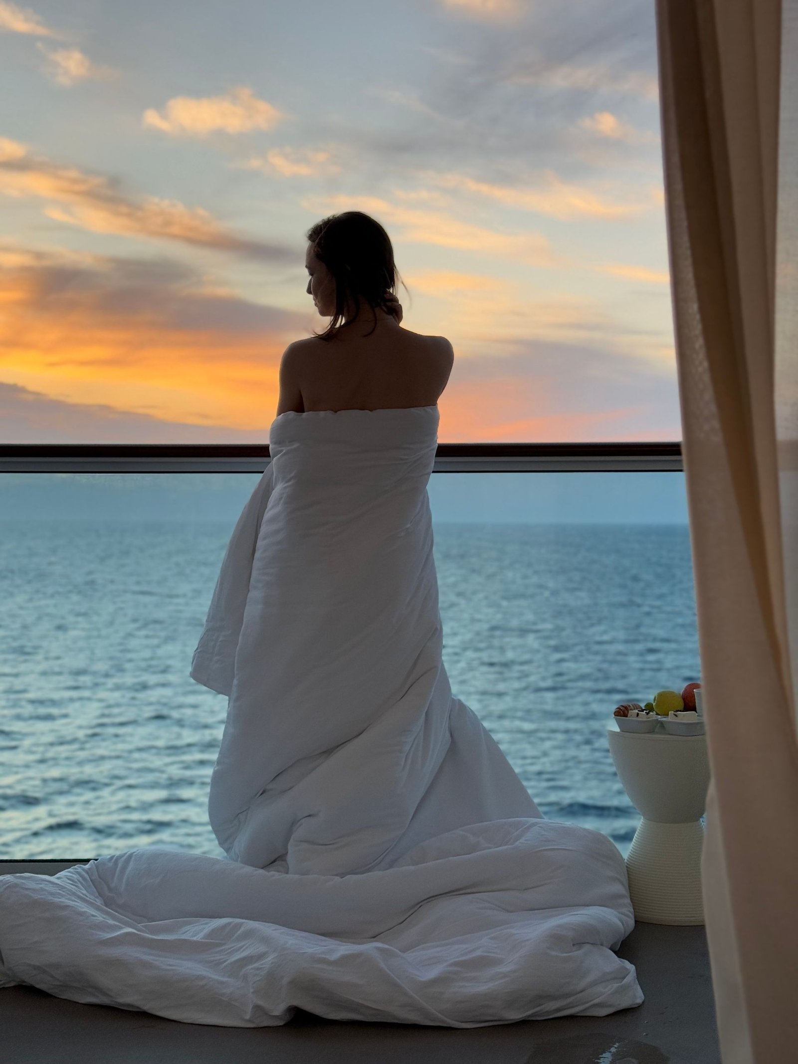 Woman wrapped in blanket enjoys a peaceful ocean view with stunning sunset backdrop.