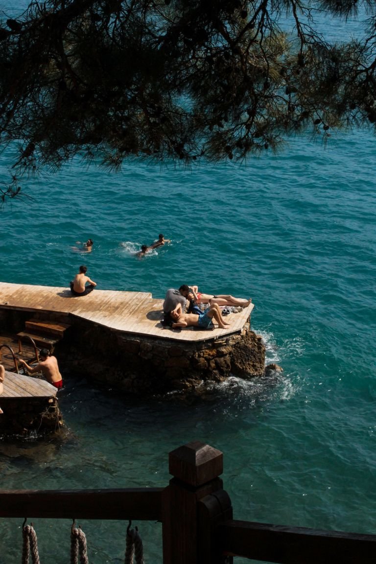 People sunbathing on a scenic deck by a clear blue sea under a tree.