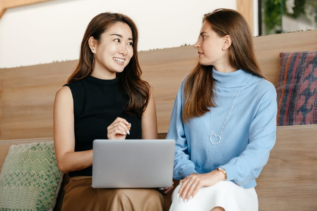 Positive young multiethnic women looking at each other while speaking and working with netbook in contemporary workplace