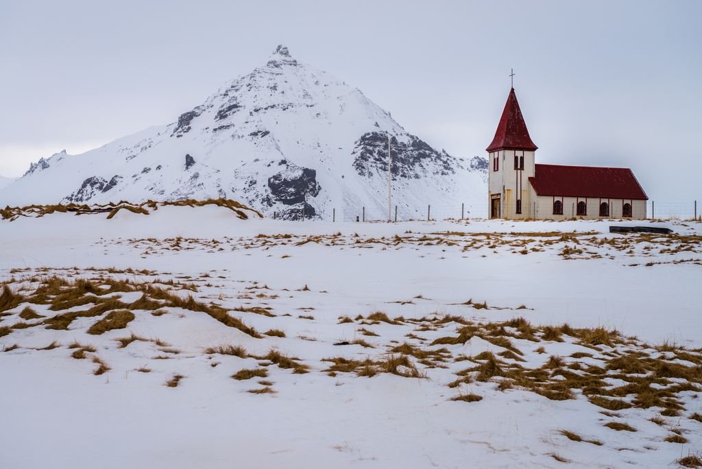 Serene winter scene of a red-roofed chapel in a snowy field with mountain backdrop.