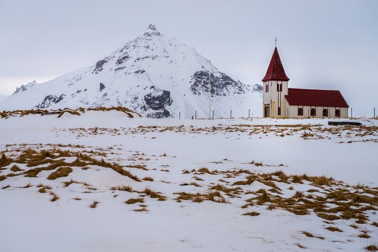 Serene winter scene of a red-roofed chapel in a snowy field with mountain backdrop.
